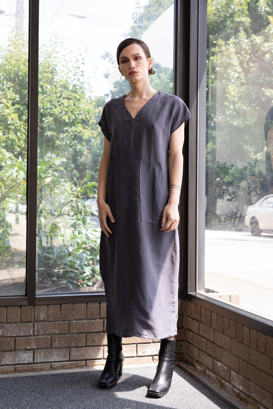 Photo of a long minimalistic dress in a stormy blue that functions made of soft Tencel linen, sleeveless with two large patch pockets on the front and a vented back.