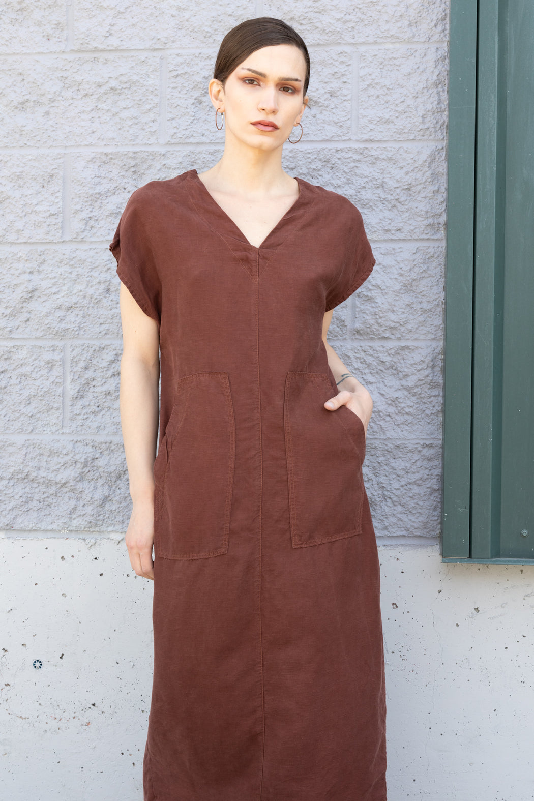 Photo of a long minimalistic dress in cinnamon brown that functions made of soft Tencel linen, sleeveless with two large patch pockets on the front and a vented back.
