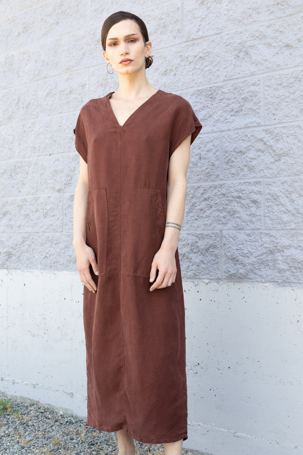 Photo of a long minimalistic dress in cinnamon brown that functions made of soft Tencel linen, sleeveless with two large patch pockets on the front and a vented back.